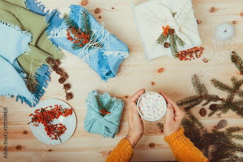 Female hands holding cup of cococa with marshmellow and just packed gift boxes wrapped in fabric and decorated with spruce branch, berries lying on wooden background. Zero waste, Sustainable Christmas photo