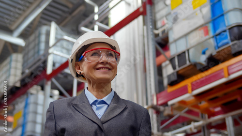 Low angle view of aged woman supervisor in safety helmet and hardhat smiling in industrial warehouse