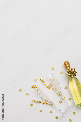 Festive white background with gold decoration , bottle of sparkling wine with two crystal glasses, shiny golden serpentine confetti and glittering snowflakes, copy space, top view