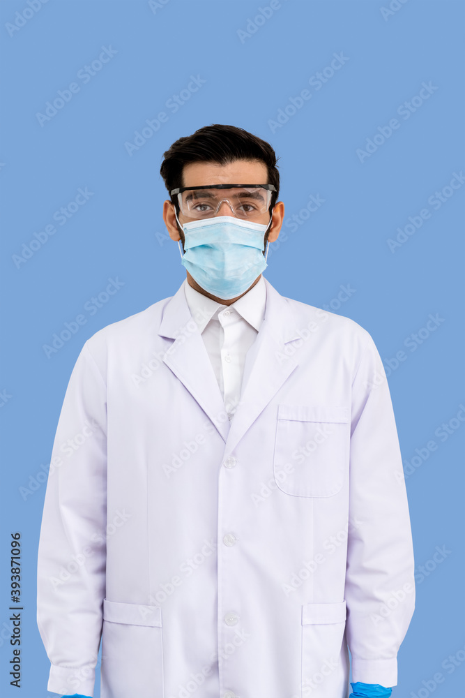 Portrait of confident male Middle Eastern Arab researcher scientist medical doctor wearing surgical mask and protection glasses standing isolated on light blue background Portrait of confident male Middle Eastern Arab researcher scientist medical doctor wearing surgical mask and protection glasses standing isolated on light blue background