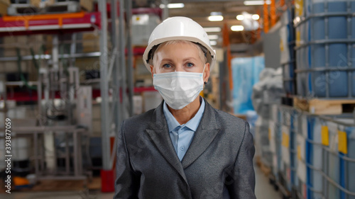 Portrait senior woman supervisor in hardhat putting on safety mask standing in industrial warehouse