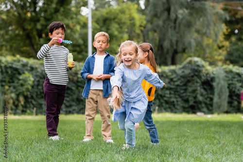 Smiling girl running on lawn near multiethnic friends with soap bubbles on blurred background