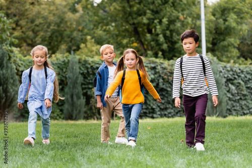 Multicultural schoolkids walking with backpacks on lawn in park