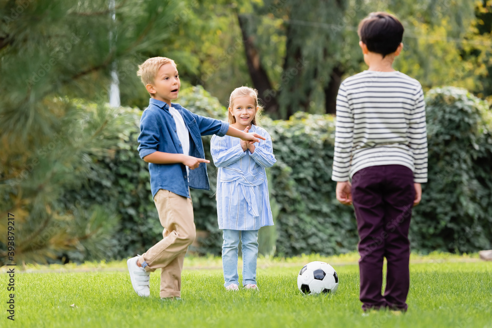 Excited boy pointing with fingers near football and friends in park Excited boy pointing with fingers near football and friends in park