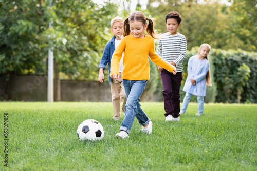 Smiling girl playing football near multiethnic friends on blurred background in park