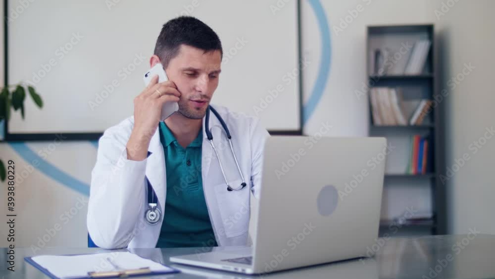 Male Doctor in White Medical Coat and Stethoscope Uses Phone to Making Remote Consulting of Patient.Telemedicine.Online Medical Consultation.Doctor Talking With Patient on Cellphone and Use a Laptop. Male Doctor in White Medical Coat and Stethoscope Uses Phone to Making Remote Consulting of Patient.Telemedicine.Online Medical Consultation.Doctor Talking With Patient on Cellphone and Use a Laptop.