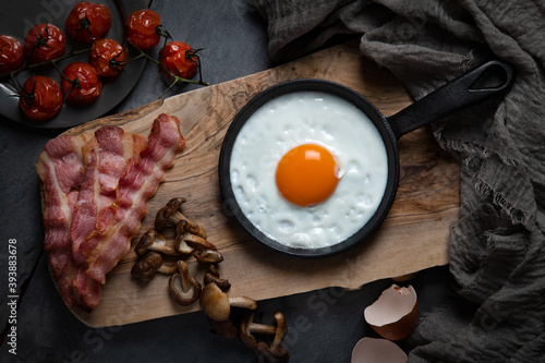 Overhead of fried egg in a pan on a wooden board, with cooked bacon, mushrooms and tomatoes arranged around, against a dark background.