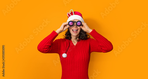 Young girl in santa claus hat on bright color background looking through binoculars. New Year and Christmas celebration concept. Woman in red hat stands having smiling holding in hand binoculars