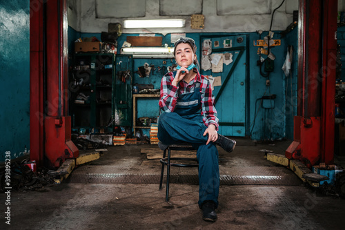 A young female pretty mechanic in a work uniform is sitting on a chair, with a medical mask on her chin. Indoor. Garage © _KUBE_