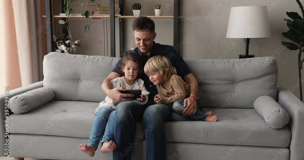 Loving father hug little daughter preschool son sit on couch with smart phone, family having fun together on internet, watch cartoons, play games online, develop kids use modern wireless tech concept Loving father hug little daughter preschool son sit on couch with smart phone, family having fun together on internet, watch cartoons, play games online, develop kids use modern wireless tech concept