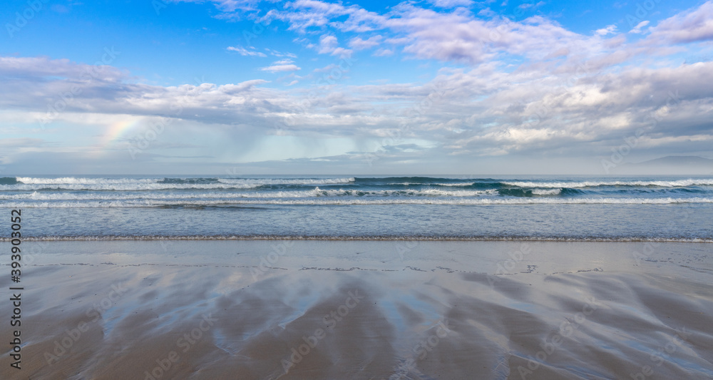 sandy beach with big waves and a small rainbow in an expressive sky