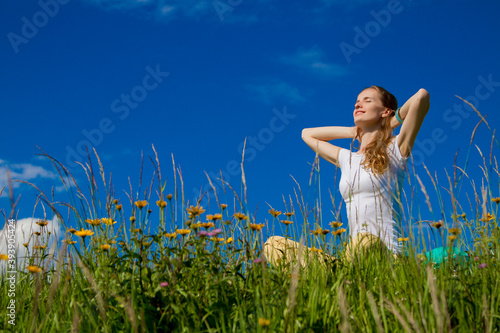 Portrait of lovely smiling woman with closed eyes sitting with raised hands and rejoicing in the sun on a meadow with wildflowers and grass against blue sky background.