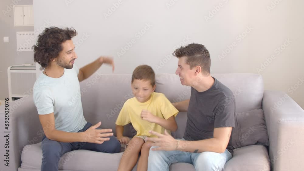 Happy joyful boy playing and dancing on couch, having fun with his two dads. Static shot, front view. Parenthood and family concept Happy joyful boy playing and dancing on couch, having fun with his two dads. Static shot, front view. Parenthood and family concept