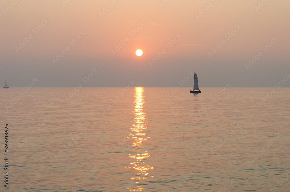 A sail boat peacefully sails at the horizon at suset time