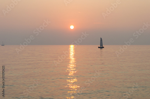 A sail boat peacefully sails at the horizon at suset time