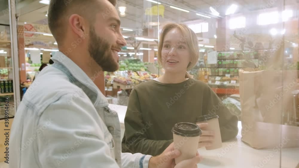 Handheld medium shot of cheerful young man and woman chatting and drinking coffee from to-go cups at market Handheld medium shot of cheerful young man and woman chatting and drinking coffee from to-go cups at market