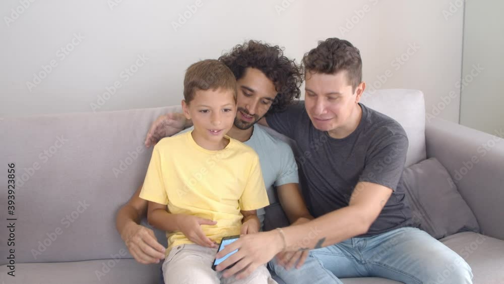 Gay parents and son spending time together at home, using and putting away smartphone, sitting on couch and smiling. Family at home and communication concept Gay parents and son spending time together at home, using and putting away smartphone, sitting on couch and smiling. Family at home and communication concept