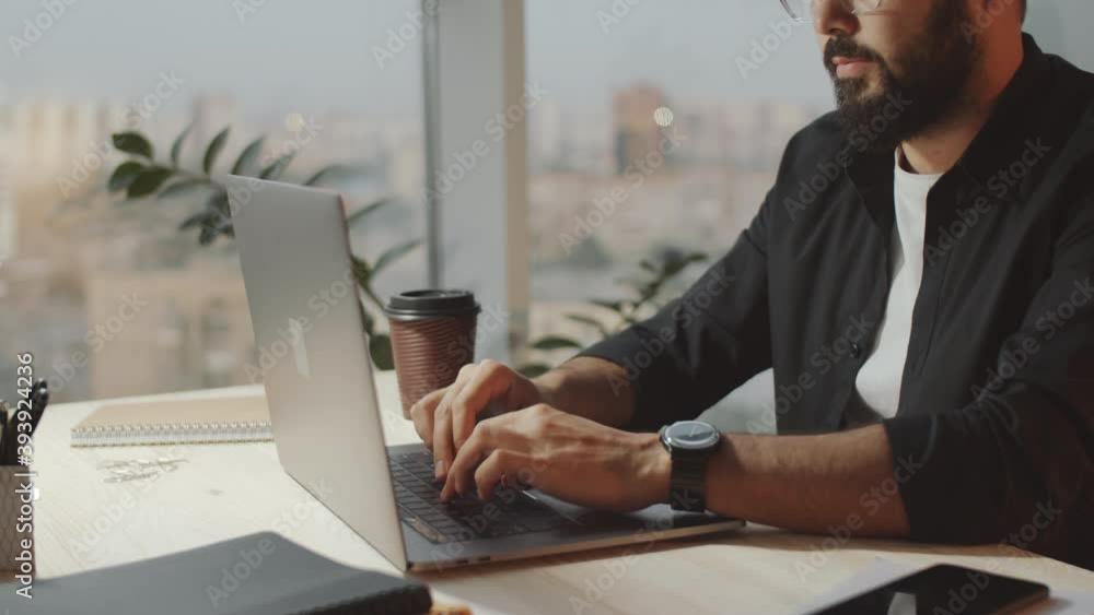 Midsection shot of young bearded businessman in casual outfit sitting at desk and typing on laptop while working in office Midsection shot of young bearded businessman in casual outfit sitting at desk and typing on laptop while working in office