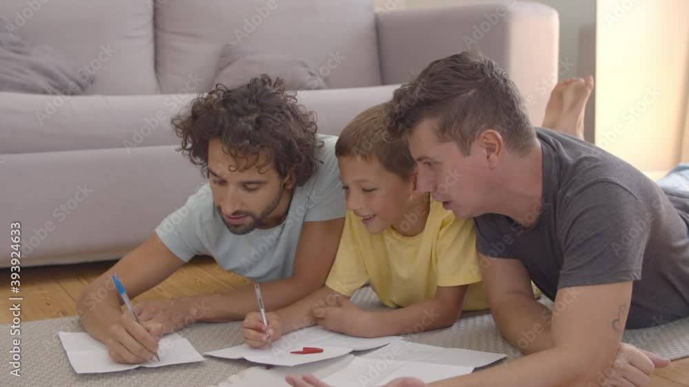Cheerful dads and son lying on floor and drawing in papers together. Static shot. Home activities and gay parents concept Cheerful dads and son lying on floor and drawing in papers together. Static shot. Home activities and gay parents concept
