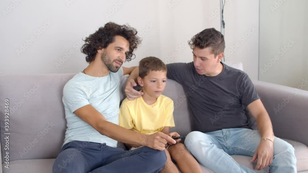 Happy homosexual parents and son sitting together in living room, talking and laughing, watching and discussing show on TV. Boy holding remote control. Family and home entertainment concept. Happy homosexual parents and son sitting together in living room, talking and laughing, watching and discussing show on TV. Boy holding remote control. Family and home entertainment concept.