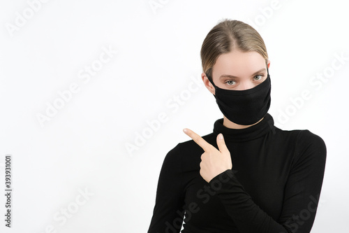 young caucasian blonde woman in disposable face protection mask points aside on free space, indoor studio photo on the white background, advertising