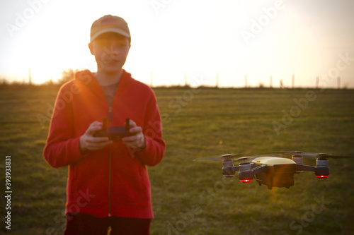 Caucasian teenage boy flying drone in a field at sunset wearing a red hoody and baseball cap
