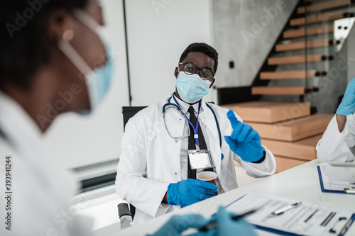 TTeam of three African-American doctors with protective face masks and gloves sitting in modern hospital and talking during business meeting. Coronavirus pandemic concept.