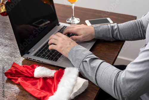 hands chatting on laptop with glass of drink and cell phone on wooden table and christmas hat and bauble on laptop screen