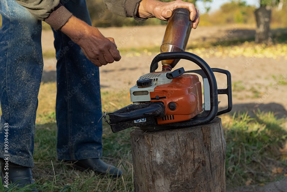  An elderly man's hands are refueling a chainsaw on a rotten tree stump.