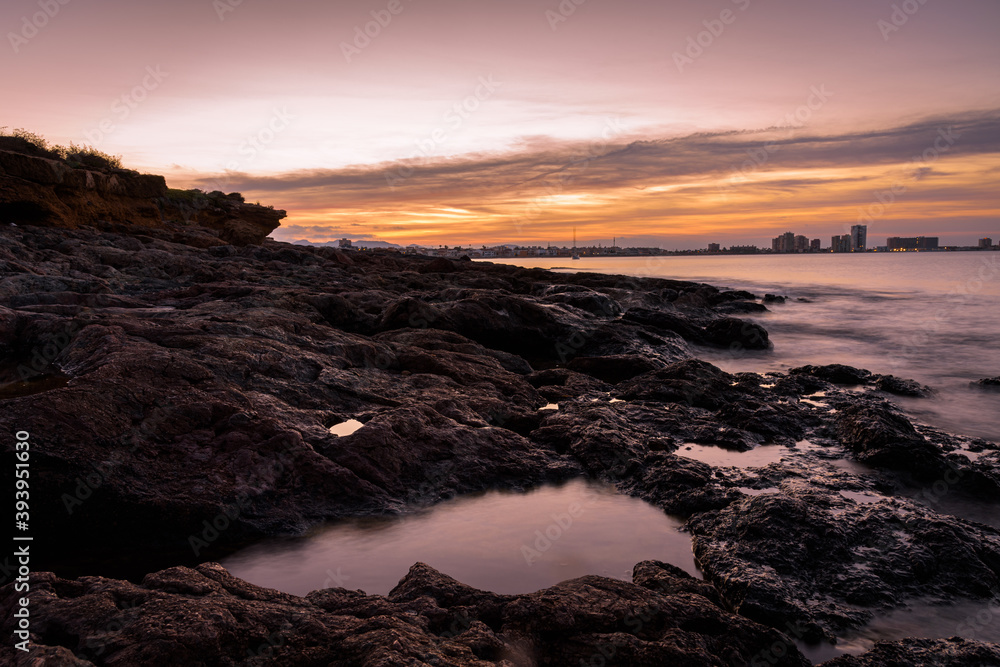 Stunning sunset from a rocky beach in Cabo de Palos with La Manga del Mar Menor skyline in the background, Murcia, Spain