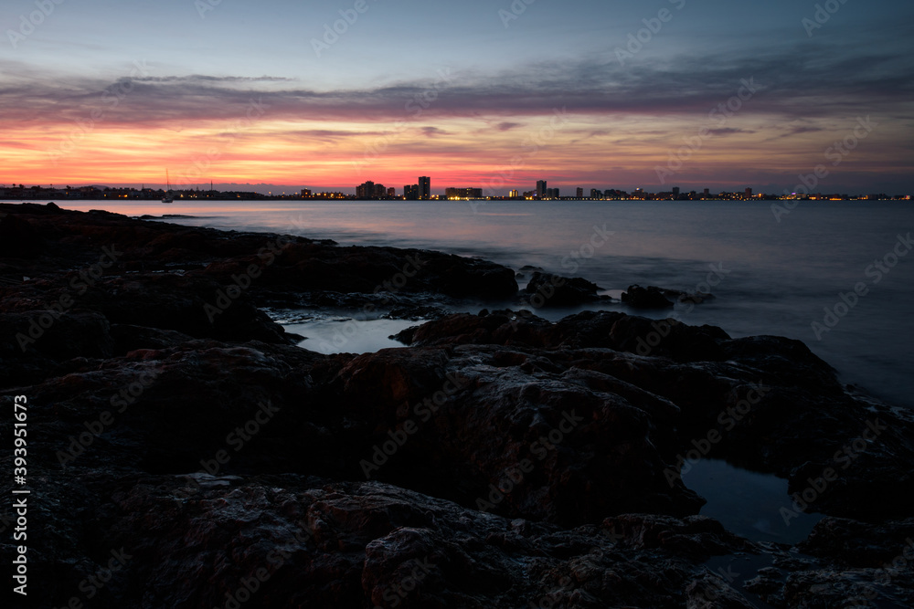 Obraz premium Stunning sunset from a rocky beach in Cabo de Palos with La Manga del Mar Menor skyline in the background, Murcia, Spain