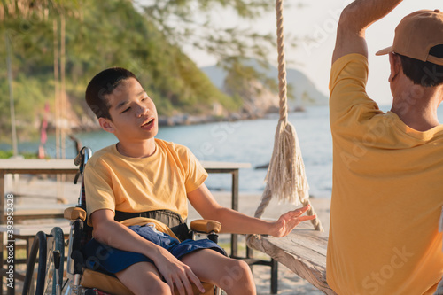 Asian special child on wheelchair smiling, playing wood swing happily on the beach with parent,Natural sea beach background,Life in the education age of disabled children,Happy disability kid concept.