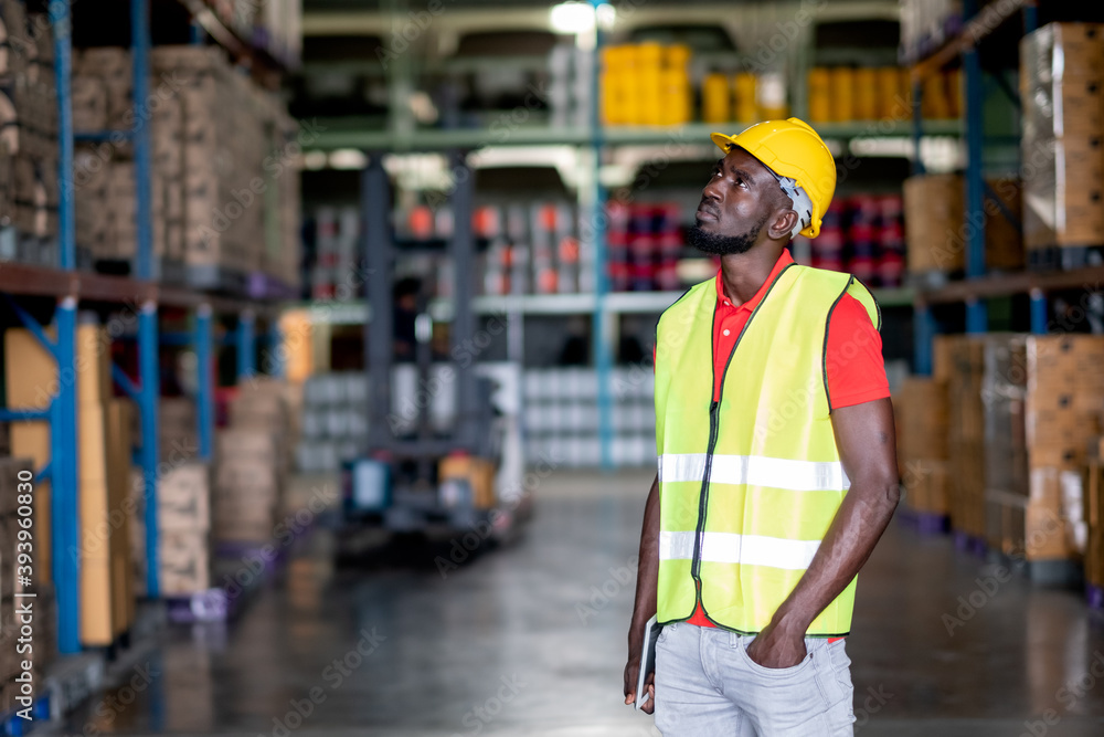 African American warehouse worker or factory man stand in front of forklift truck and look to upper left in workplace area. Concept of good management of staff work in industrial business.