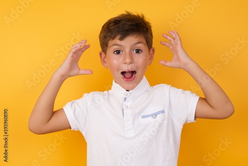 little cute Caucasian boy kid wearing white t-shirt against yellow wall looks with excitement at camera, keeps hands raised over head, notices something unexpected, reacts on sudden news.