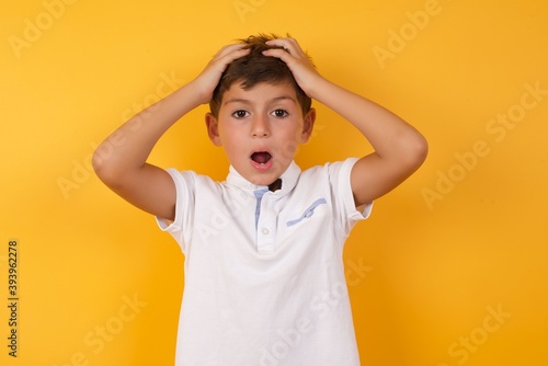 Horrible, stress, shock. Portrait emotional crazy little cute Caucasian boy kid wearing white t-shirt against yellow wall clasping head in hands. Emotions, facial expression concept.