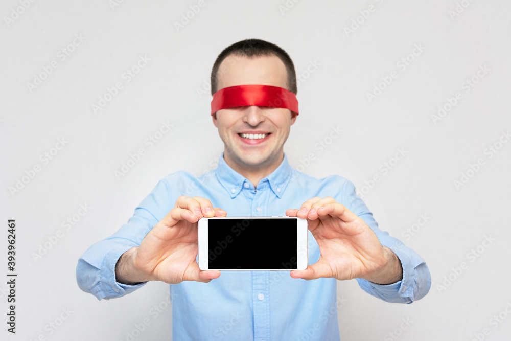 Blindfolded man shows the screen of the phone and smiles, portrait, white background, copy space Blindfolded man shows the screen of the phone and smiles, portrait, white background, copy space