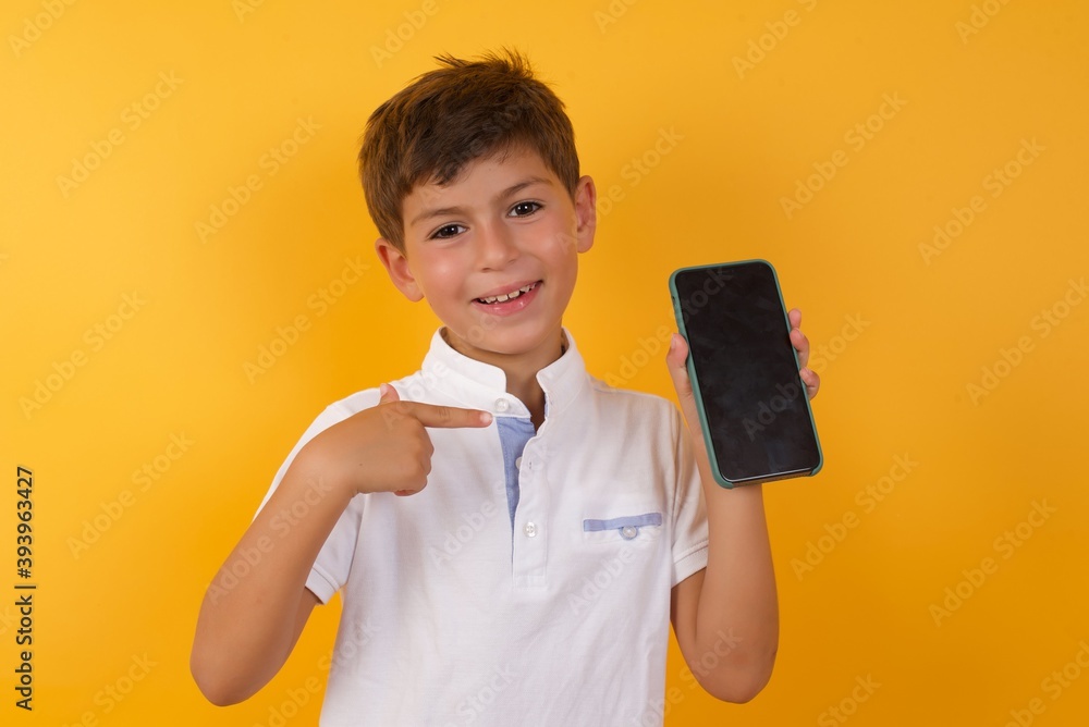 Excited little cute boy kid wearing white t-shirt against yellow wall holding and pointing with finger at smartphone with blank screen. Advertisement concept. Excited little cute boy kid wearing white t-shirt against yellow wall holding and pointing with finger at smartphone with blank screen. Advertisement concept.