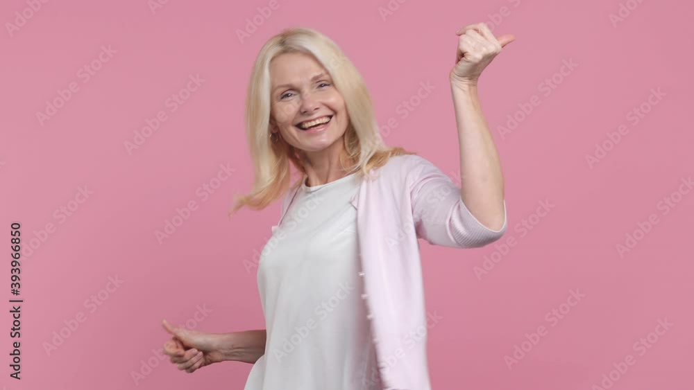 Smiling cute elderly gray-haired blond woman lady 40s 50s years old in white t-shirt dancing fooling around having fun expressive gesticulating hands isolated on pastel pink background studio portrait