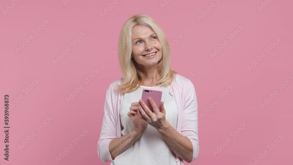Smiling beautiful elderly gray-haired woman lady 40s 50s years old in white blank casual t-shirt hold in hand using mobile cell phone chatting isolated on pastel pink color background studio portrait Smiling beautiful elderly gray-haired woman lady 40s 50s years old in white blank casual t-shirt hold in hand using mobile cell phone chatting isolated on pastel pink color background studio portrait