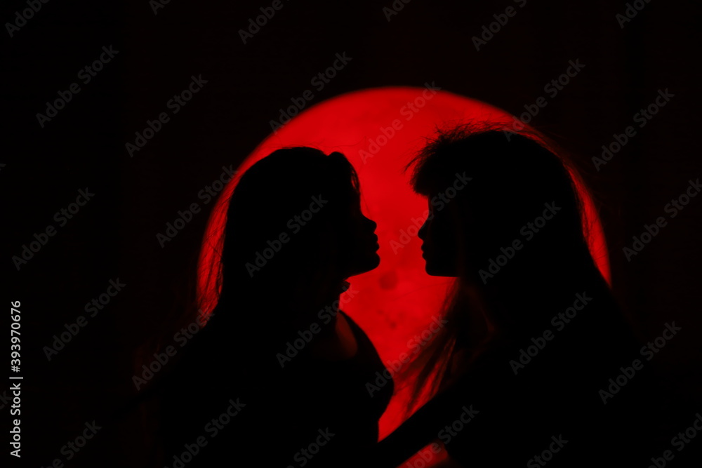 A silhouette of two female dolls standing very close to each other in front of a circular red light source in the dark	