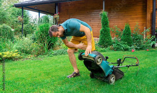 Electric lawn mower breakdown. Malfunctioning garden tools. The man overturned the grass collector lawnmower to see what was causing the breakdown. An unexpected problem that slows down the process.