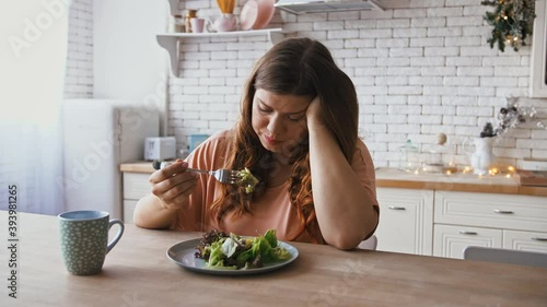 Boring diet. Depressed overweight woman eating untasty healthy salad at kitchen photo