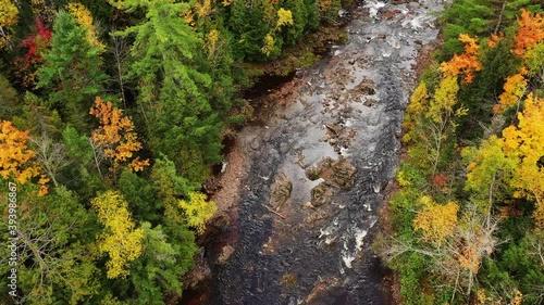 Beautiful autumn aerial view flying down and following the Montreal River heading over Saxon Falls towards the power house and footbridge with rock formations and colorful fall foliage and evergreens.