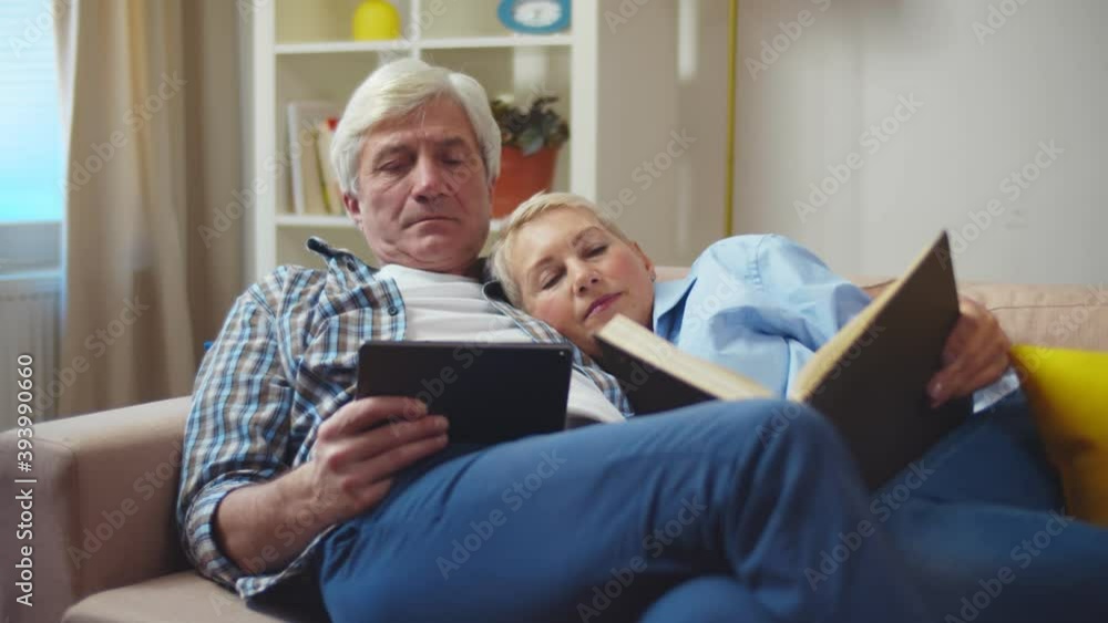 Senior couple reading book and using tablet on couch in living room