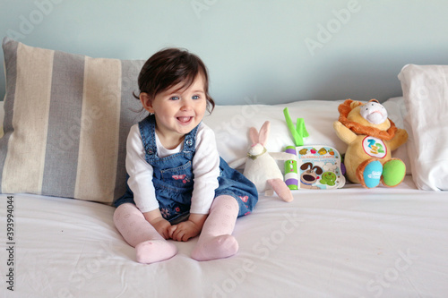 Happy smiling baby girl in denim dungarees sitting on a bed with soft toys