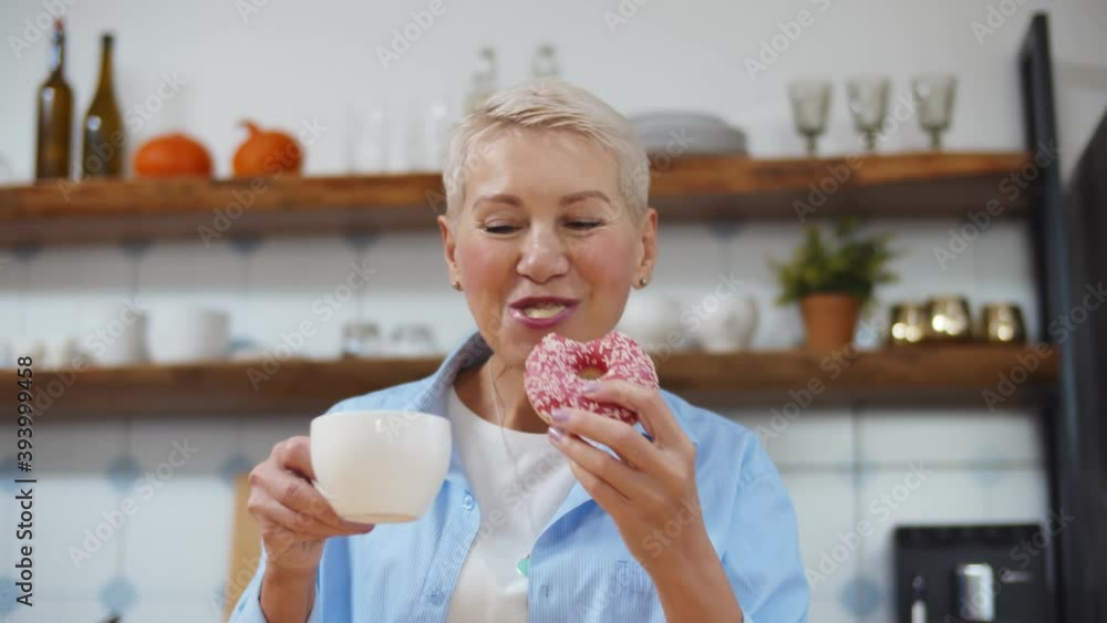Smiling senior woman drinking coffee and eating delicious glazed donut at home