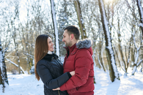 Young couple in love, enjoying winter weather outside in park forest. Brunette man in red jacket and woman in black, hugging, looking at each other. Christmas and new year holidays joy. Romantic date.