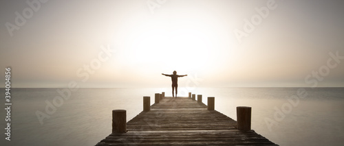 Fotografie, Obraz Person stands with open arms on the pier at sunrise