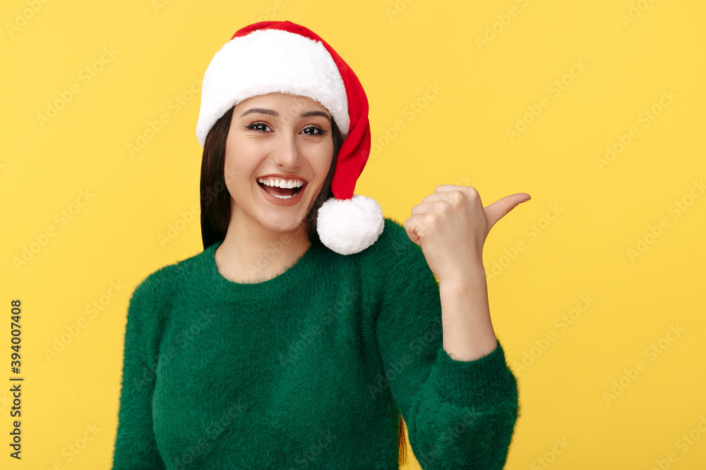 Positive young brunette woman wear santa claus hat and green pullover pointing thumb away over yellow background. Positive young brunette woman wear santa claus hat and green pullover pointing thumb away over yellow background.