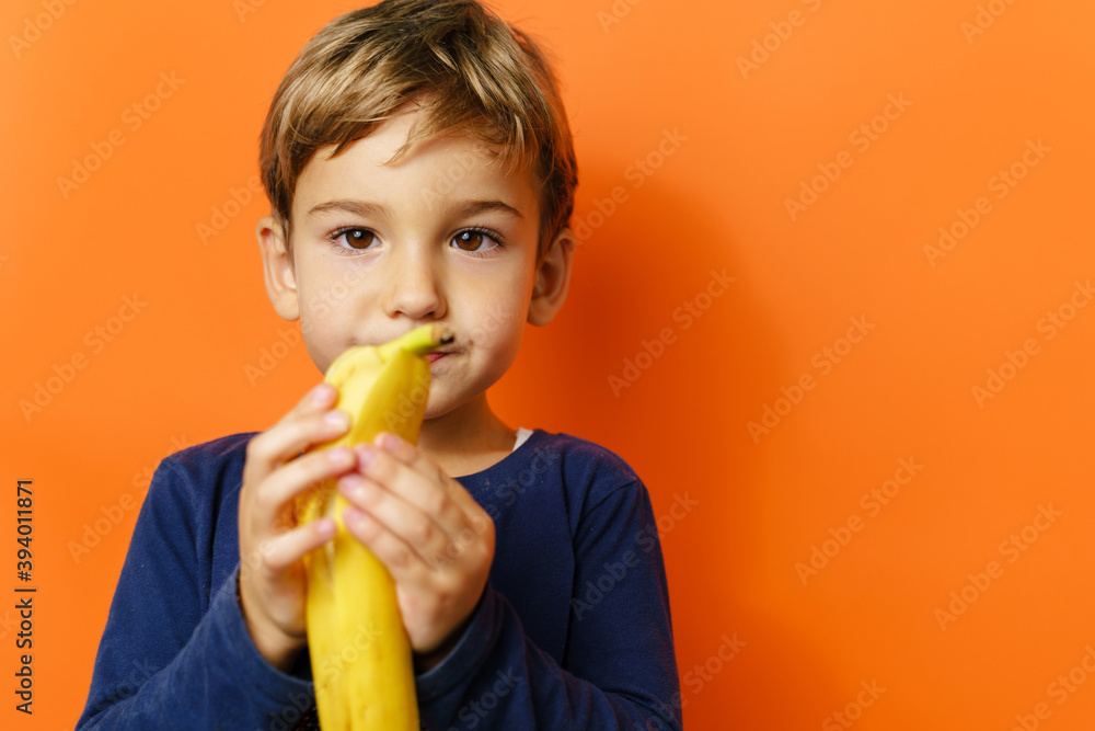 Small caucasian boy eating banana in front of orange background wall Small caucasian boy eating banana in front of orange background wall